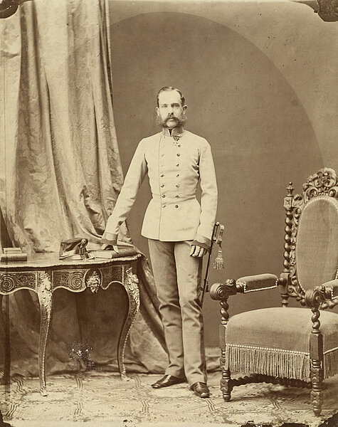 Schwarzweißfotografie von Kaiser Franz Joseph I als junger Mann zwischen einem eleganten Schreibtisch und einem Sessel