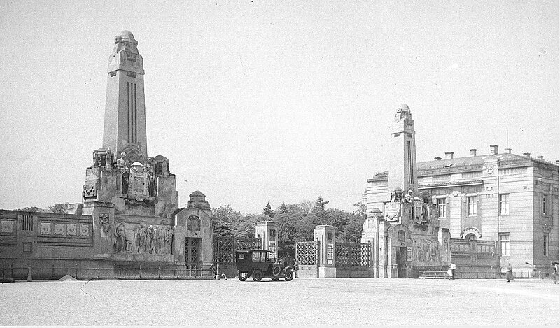 Schwarz-weißes Foto von Friedhofstor mit zwei Statuen links und rechts, davor ein altes Auto.