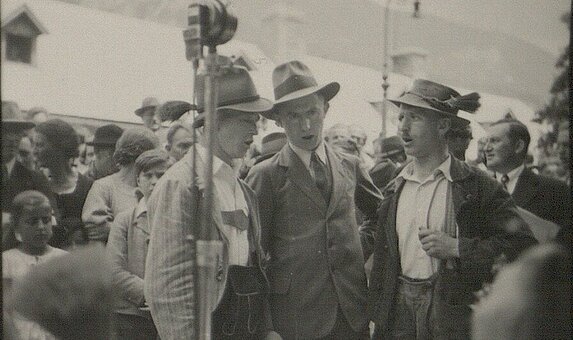 Schwarz-weißes Foto von drei Personen in Tracht an einem Mikro stehend.