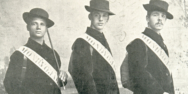 Schwarz-weißes Foto von drei Personen in Anzügen und Hüten mit einer weißen Schärpe.