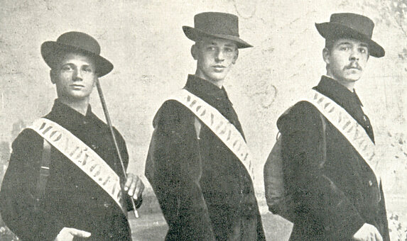 Schwarz-weißes Foto von drei Personen in Anzügen und Hüten mit einer weißen Schärpe.